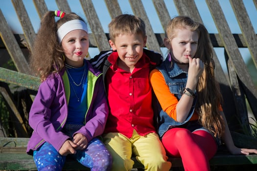 Funny little children girls with bright makeup dressed in the style of the nineties and a boy in a red shirt are sitting on a bench by a skewed fence. Russian village children. photo