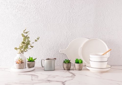 white ceramic utensils and indoor flowers in ceramic mini pots on the kitchen countertop made of white marble. grey cement wall. a copy space. photo