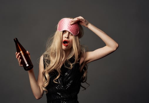 Drunk woman with a bottle of alcohol in her hand on a gray background and a mask for sleeping photo