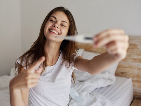 cheerful woman sitting on the bed with a thermometer in her hands, positive result photo