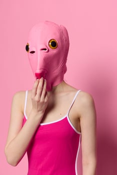 A funny woman wearing a pink fish mask on her head put her fingers in her mouth with pink clothes on a pink background. The concept of modern art photography photo