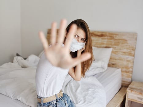 red-haired woman in a medical mask is covered by her hands from the camera while sitting on the bed indoors photo