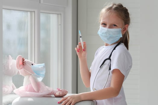 A cute little girl in a medical mask holds a syringe while vaccinating a toy against the Covid-19 coronavirus. Smart child playing hospital game pretending to be doctor, veterinarian photo