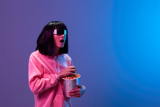 Shocked adorable brunet woman in pink hoodie trendy specular sunglasses with popcorn open mouth look aside posing isolated in blue violet color light background. Neon party Cinema concept. Copy space photo