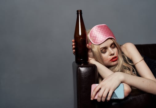 Drunk blonde woman with a bottle of beer sleeps on the sofa indoors on a gray background and a sleep mask on her head photo