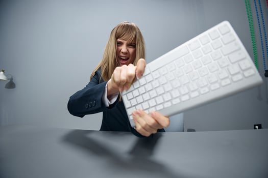 Crazy irritated angry millennial blonde businesswoman worker in blue jacket throws keyboard and screaming at camera in gray modern office. People Emotions Business Concept. Copy space, wide angle photo