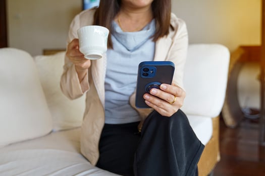 Asian woman sitting on sofa sipping coffee and watching online news on mobile phone in the morning before going to work. Soft and selective focus. photo