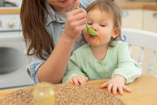 Happy family at home. Mother feeding her baby girl from spoon in kitchen. Little toddler child with messy funny face eats healthy food at home. Young woman mom giving food to kid daughter. photo