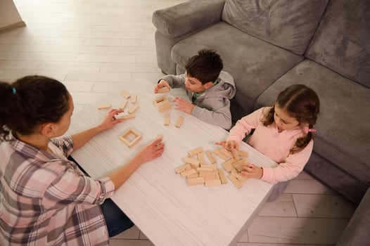 Overhead view of a mom and kids playing board games, building wooden structures, constructions on the table in home living room. Family pastime and educational leisure concept photo