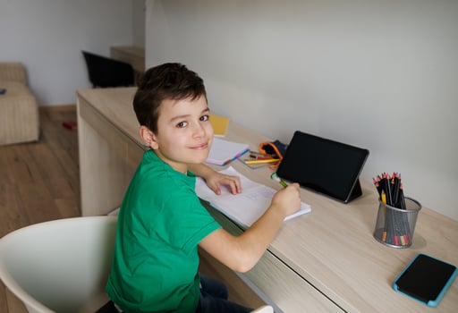 Handsome Caucasian 9 years old school boy sits at wooden desk in front of a digital gadget and doing his homework, cutely smiles looking at camera. Education and school concept. Online learning photo