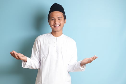 Portrait of Asian muslim man in white koko shirt with skullcap pulling hands towards camera, inviting someone to come inside, welcoming gesture. Isolated image on blue background photo