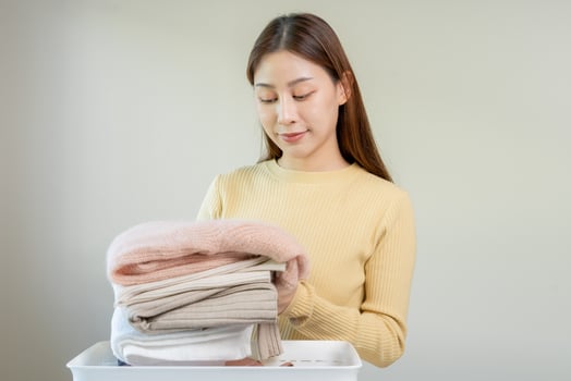 Chore of housewife, smile asian young woman folding many freshly shirts put the cloth in the basket after washing clean clothes and drying, making household working in room at home, maid laundry tidy. photo