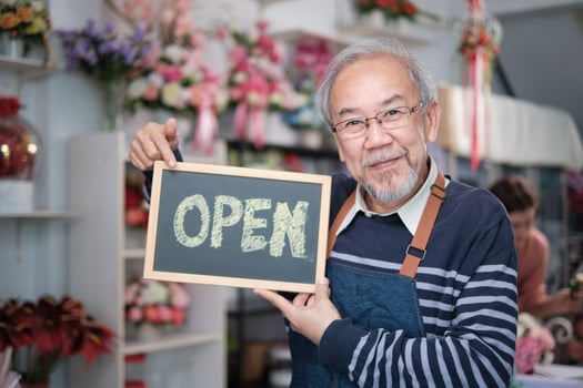 One senior male florist owner in apron shows open sign board in bright flower shop store with smile and looks at camera, small business opening, occupation retirement, happy elderly SME entrepreneur. photo
