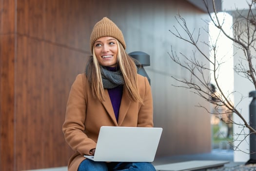 beautiful young woman wearing autumn coat using laptop while sitting on a bench, drinking takeaway coffee cup photo