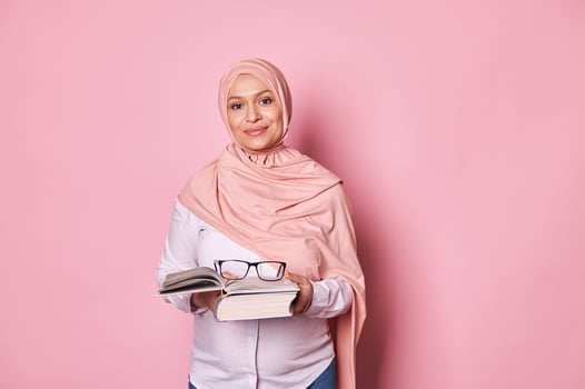 Confident Muslim woman educator in pink hijab, a school teacher holding eyeglasses and studying book, pink background photo