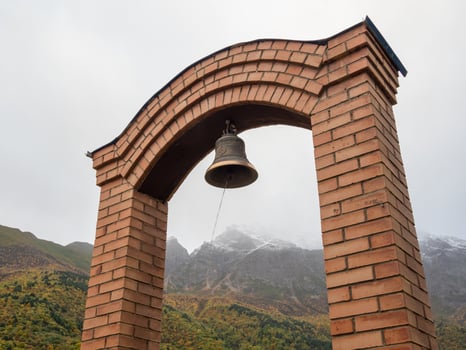 Stock photo of ancient copper or bronze bell hanging on a stone frame at Chapel in honor of the dead on foggy mountain background. Picture captured at Irafsky district, Stur-Digora village. Russia.
