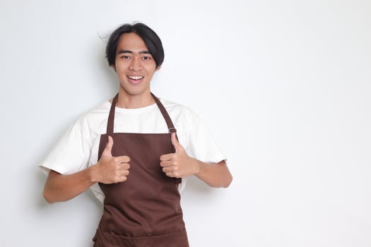 Portrait of attractive Asian barista man in brown apron showing thumb up hand gesture. Advertising concept. Isolated image on white background photo