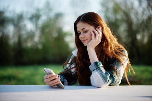 A woman with curly red hair in a plaid shirt sits outdoors in the park with a cell phone in her hands and looks away. Freelance work in the park photo