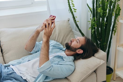 A man with a beard lies on the couch during the day at home and looks at his phone relaxing on his day off, a man gambling on the stock market online on his phone photo