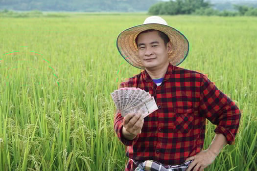 Asian farmer man is at paddy field, wears hat and red plaid shirt, hold Thai banknote money. Concept , Farmer happy to get profit, income, agriculture supporting money. photo