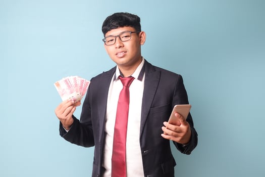 Portrait of young Asian business man in casual suit looking at camera with slight head tilt while holding phone thousand rupiahs. Isolated image on blue background photo