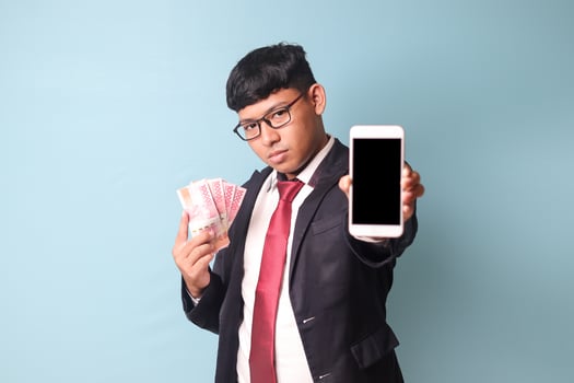 Portrait of young Asian business man in casual suit showing phone and holding thousand rupiahs looking at camera with serious face. Isolated image on blue background photo