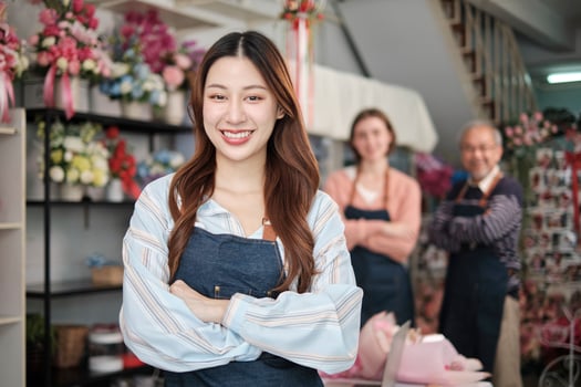 Beautiful young Asian female florist worker in apron, arms crossed, smiles and looks at camera in front of colleagues team in colorful flower shop, small business occupation, happy SME entrepreneur. photo