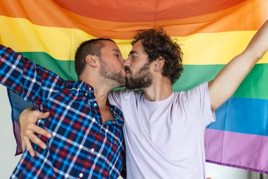 Two young gay lovers kissing each other affectionately. Two young male lovers standing together against a pride flag. Affectionate young gay couple sharing a romantic moment together. photo