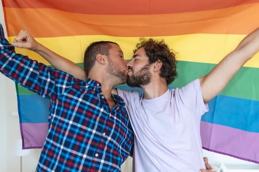Two young gay lovers kissing each other affectionately. Two young male lovers standing together against a pride flag. Affectionate young gay couple sharing a romantic moment together. photo