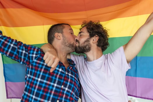Two young gay lovers kissing each other affectionately. Two young male lovers standing together against a pride flag. Affectionate young gay couple sharing a romantic moment together. photo