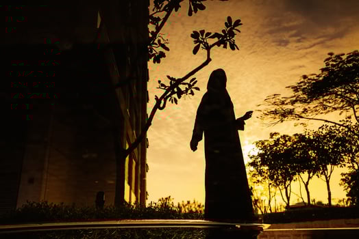 a young woman wearing a hijab and jilbab standing holding a phone near a water body at dusk photo