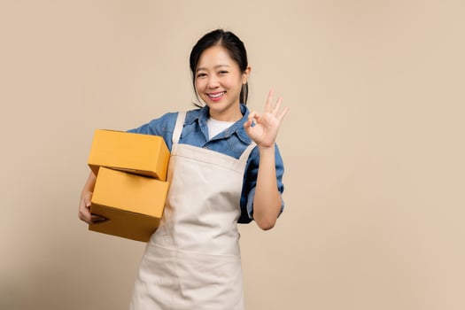 Young Asian woman wearing brown apron and holding parcel box and sitting on isolated over the light brown background , SME Online marketing packing box delivery and isolated concept photo