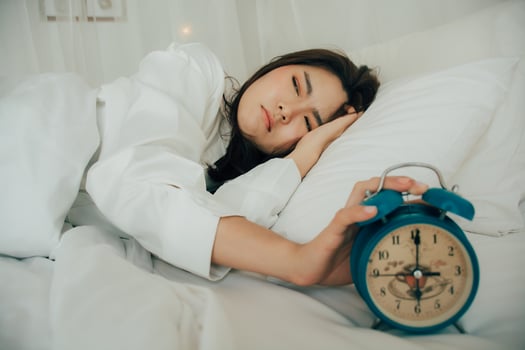 Asian beautiful woman in white pajamas turning off alarm clock while sleepy in bed at her bedroom of house or hotel on holidays. Girl is having trouble wake up late in morning. Unhealthy sleep. photo