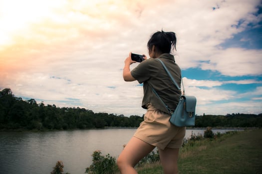 Young asian girl to travel the holidays. She was wearing a green shirt - shoulder bag, looking at the sunset and using mobile phone to take pictures of river and mountains or video call feel relaxed. photo