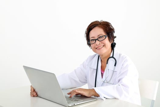 Elderly asian Chinese female doctor on white background wearing white apron stethoscope look at camera typing working at laptop notebook pc photo