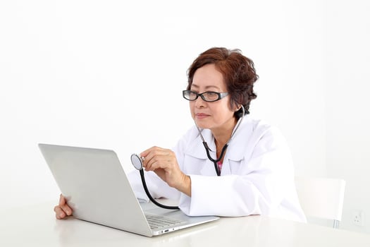 Elderly asian Chinese female doctor on white background wearing white apron checking diagnostics on notebook laptop pc with stethoscope photo