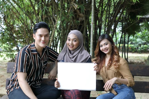 Young Asian Malay man woman headscarf outdoor green sit on park bench paint draw hold white board copy space text photo