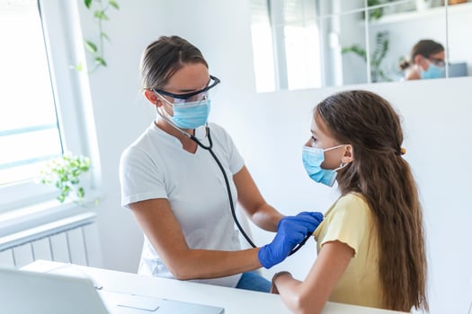 Adorable girl is sitting at her doctor's office. Her doctor is using a stethoscope to listen to her chest. Both her and her doctor are wearing a face mask to prevent the spread of germs. photo