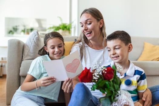 Young mother with a bouquet of roses laughs, hugging her son, and cheerful girl with a card and roses congratulates mom during holiday celebration in kitchen at home photo