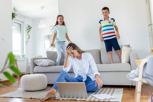 Tired young mother sitting on the floor and working with laptop and documents while little kids and jumping on the sofa and having fun and making noise photo