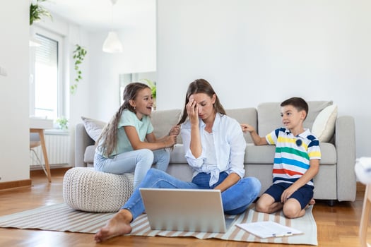 Small mischievous kids noising and distracting mother freelancer trying to concentrate on laptop and squeezing head working at table in light living room photo