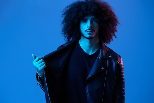 Portrait of fashion man with curly hair on blue background multinational, colored light, black leather jacket trend, modern concept. photo