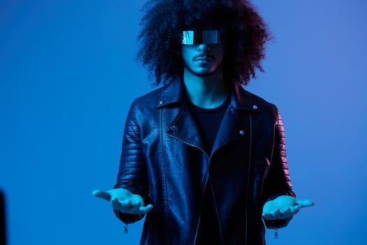 Portrait of fashion man with curly hair with stylish glasses on blue background multinational, colored light, black leather jacket trend, modern concept. photo
