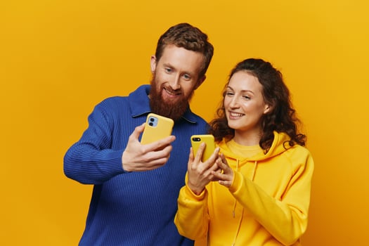 Man and woman couple smiling merrily with phone in hand social media viewing photos and videos, on yellow background, symbols signs and hand gestures, family freelancers.