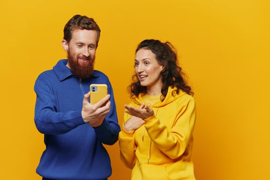 Man and woman couple smiling merrily with phone in hand social media viewing photos and videos, on yellow background, symbols signs and hand gestures, family freelancers.