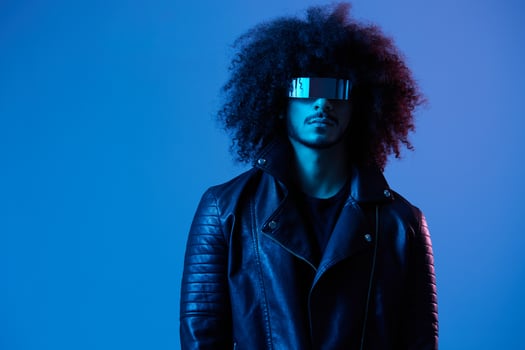 Portrait of fashion man with curly hair with stylish glasses on blue background multinational, colored light, black leather jacket trend, modern concept. photo