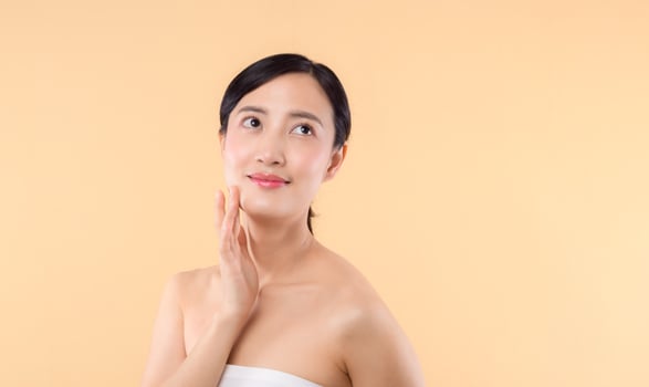 Beautiful girl asian model touching fresh glowing hydrated facial skin on beige background closeup. Beauty face young woman with natural makeup and healthy skin portrait. Skin care concept photo