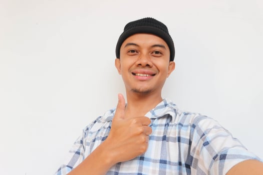 Happy adult man wearing pajamas and beanie cap smiling while showing thumb up and looking at camera, OK sign. Isolated on white background photo