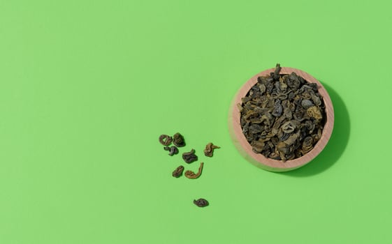 Dry rolled green tea in a wooden bowl on a green background, top view photo