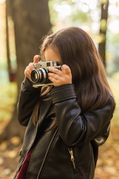 A little girl take a photo with old retro camera in autumn nature. Leisure and hobby concept.